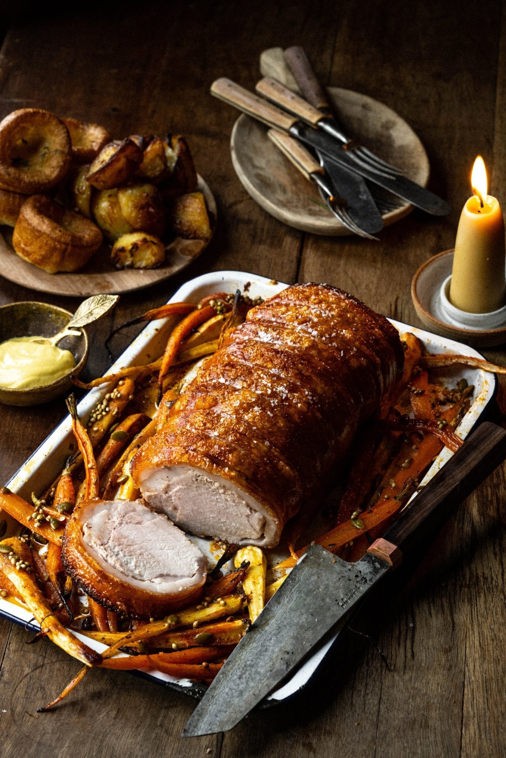 A whole roast pork loin with crisp golden crackling, two slices carved, resting in an enamel tray of roasted carrots and parsnips