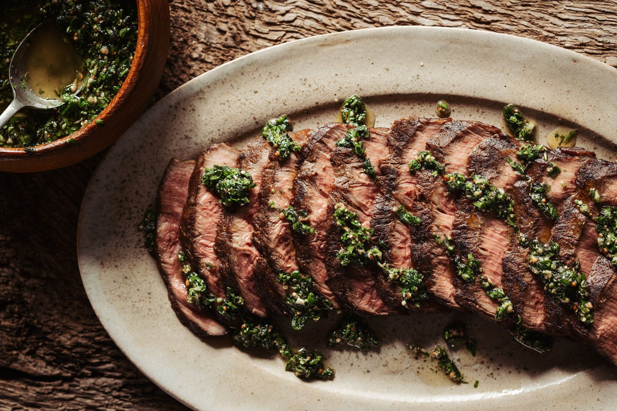 Sliced steak with green herb sauce on a white plate, wooden background