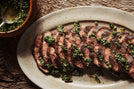 Sliced steak with green herb sauce on a white plate, wooden background